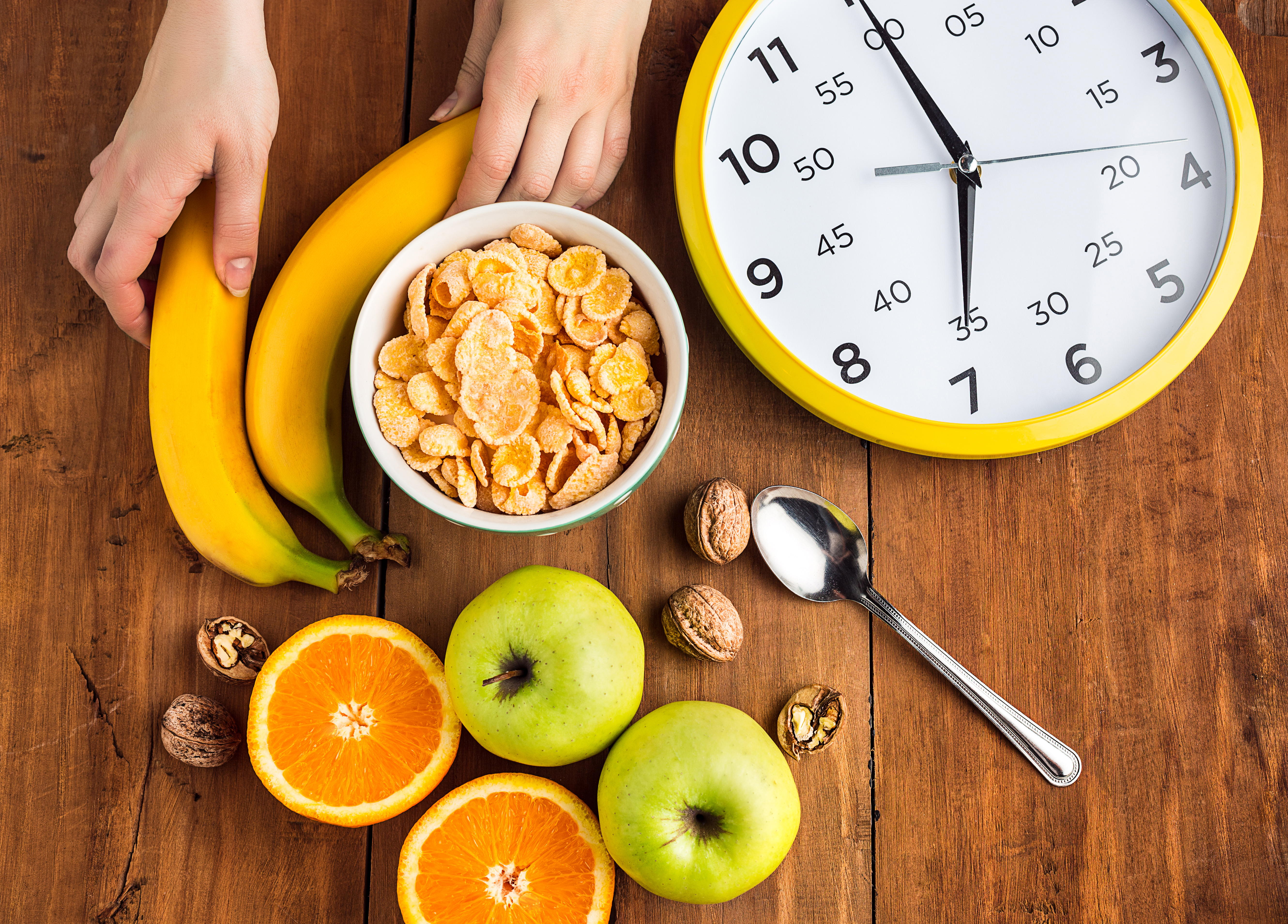 Fruits frais comme des bananes, des pommes et des oranges avec un bol de c&eacute;r&eacute;ales, des noix et une horloge jaune sur une table en bois, illustrant le moment de rompre le je&ucirc;ne intermittent avec un repas sain