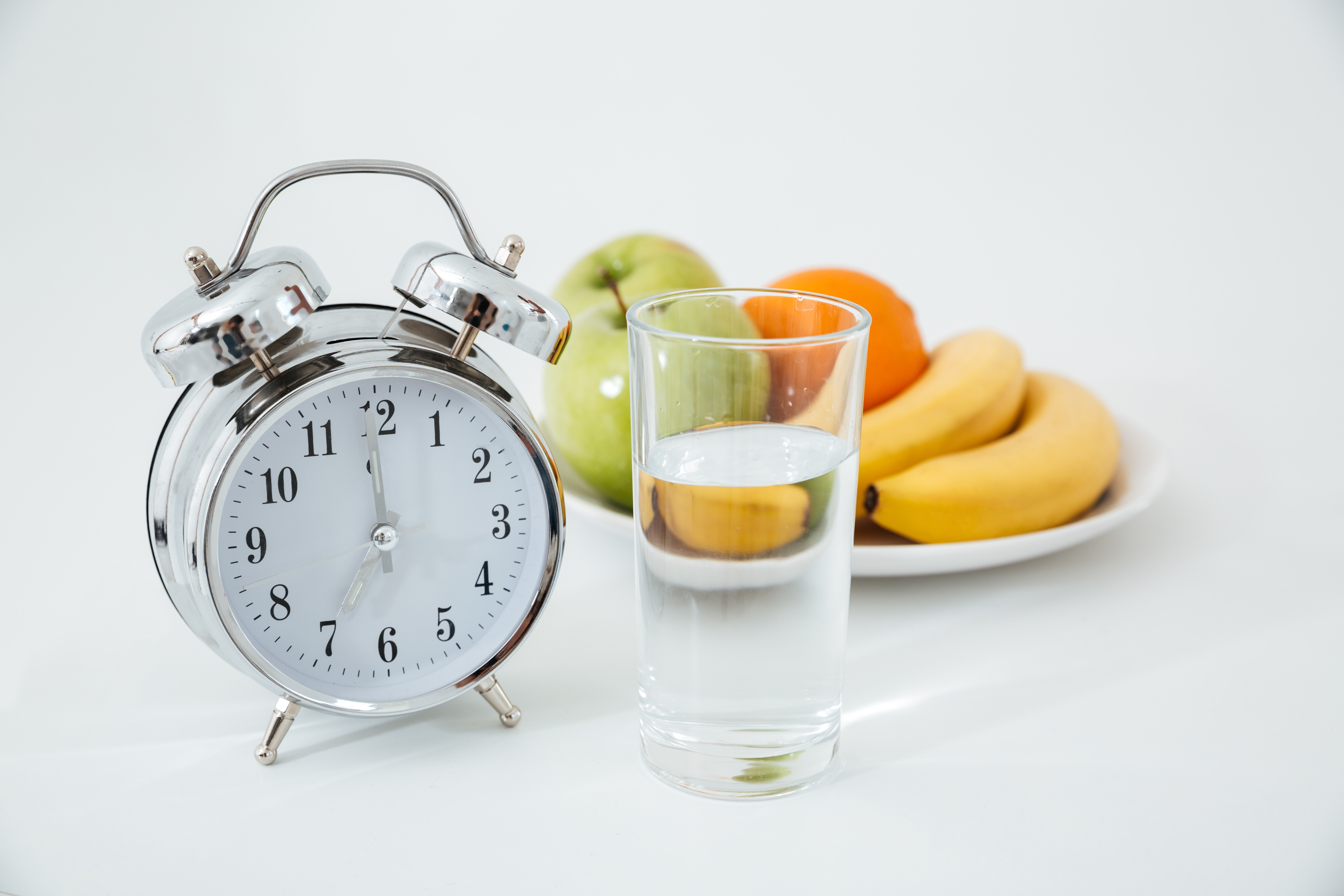 R&eacute;veil pos&eacute; &agrave; c&ocirc;t&eacute; d&rsquo;un verre d&rsquo;eau et d&rsquo;une assiette de fruits frais comme des bananes, des pommes et des oranges, illustrant le concept du je&ucirc;ne intermittent et d&rsquo;une alimentation saine