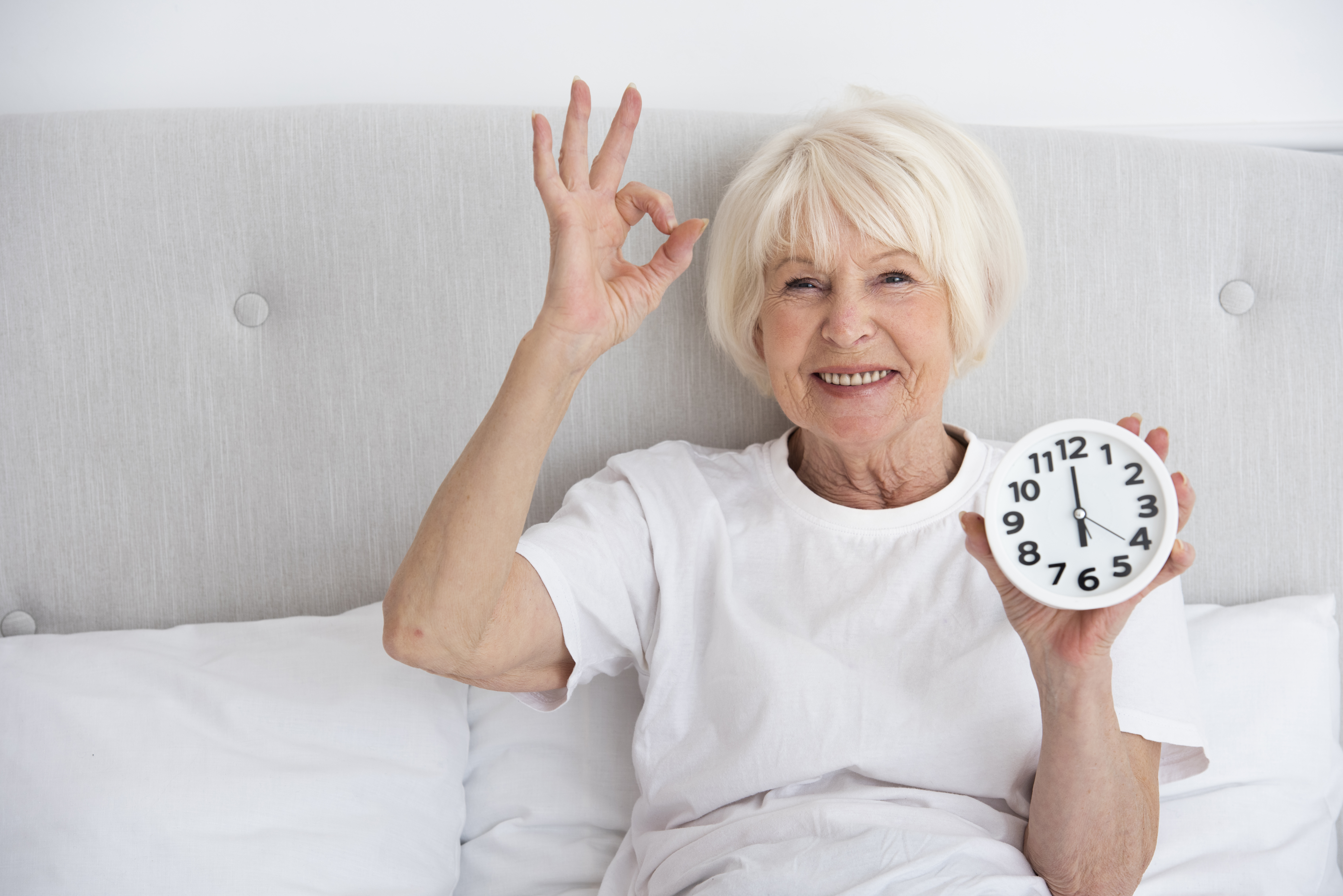 Femme &acirc;g&eacute;e souriante assise sur un lit en v&ecirc;tements blancs, tenant une horloge et faisant le geste &ldquo;OK&rdquo; de la main, repr&eacute;sentant le bien-&ecirc;tre, le vieillissement en bonne sant&eacute; et la gestion du temps dans le cadre du je&ucirc;ne intermittent